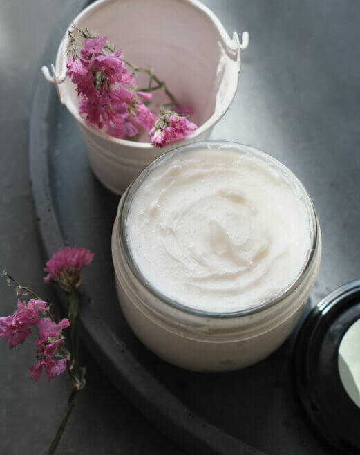 Hautpflegecreme in einem Glas mit getrockneten Blumen und leerem Behälter auf einem Tisch.